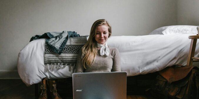 woman sitting beside a bed while using a laptop